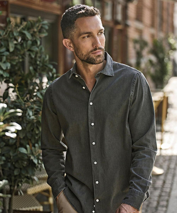 A man with short hair and a trimmed beard wears a Tee Jays Denim shirt (4004) and stands outdoors near greenery and a building, looking to his left.