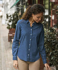 A woman with curly hair wears a Tee Jays Womens denim shirt (4005) and beige trousers, standing outdoors by green leafy plants and looking down.