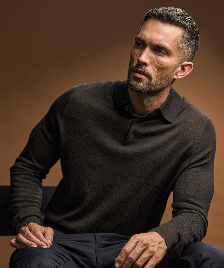 A man with short dark hair and a trimmed beard is seated on a chair against a brown background, wearing the Tee Jays Polo knit (6002) long-sleeve collared shirt and dark trousers, looking slightly to the side.