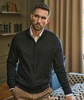 A man with short hair and a trimmed beard is wearing the Tee Jays Full-zip knit (6012) over a white collared shirt and tan trousers. He stands indoors, one hand in his pocket, beside a sofa and bookshelves.