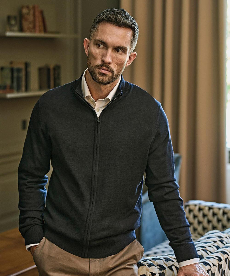 A man with short hair and a trimmed beard is wearing the Tee Jays Full-zip knit (6012) over a white collared shirt and tan trousers. He stands indoors, one hand in his pocket, beside a sofa and bookshelves.