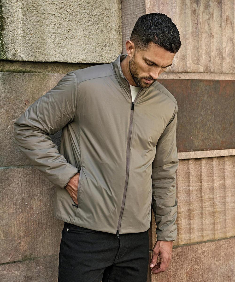 A man stands outdoors against a stone wall, wearing the Tee Jays PureLite jacket (9690) in light brown and black trousers, with one hand in his pocket and looking down.