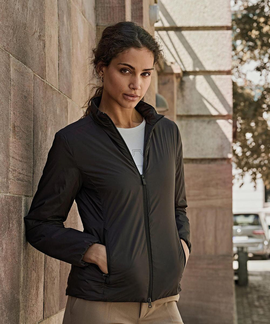 A woman with curly hair pulled back stands outdoors against a stone wall, hands in pockets, wearing the Tee Jays Womens PureLite jacket (9691). Trees and a parked car are in the background as she looks directly at the camera.