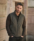 A man with short hair and a trimmed beard stands outside against a stone wall, wearing a Tee Jays PureLite bodywarmer (9692) in olive green over a dark brown long-sleeve shirt, with his hands in his pockets.