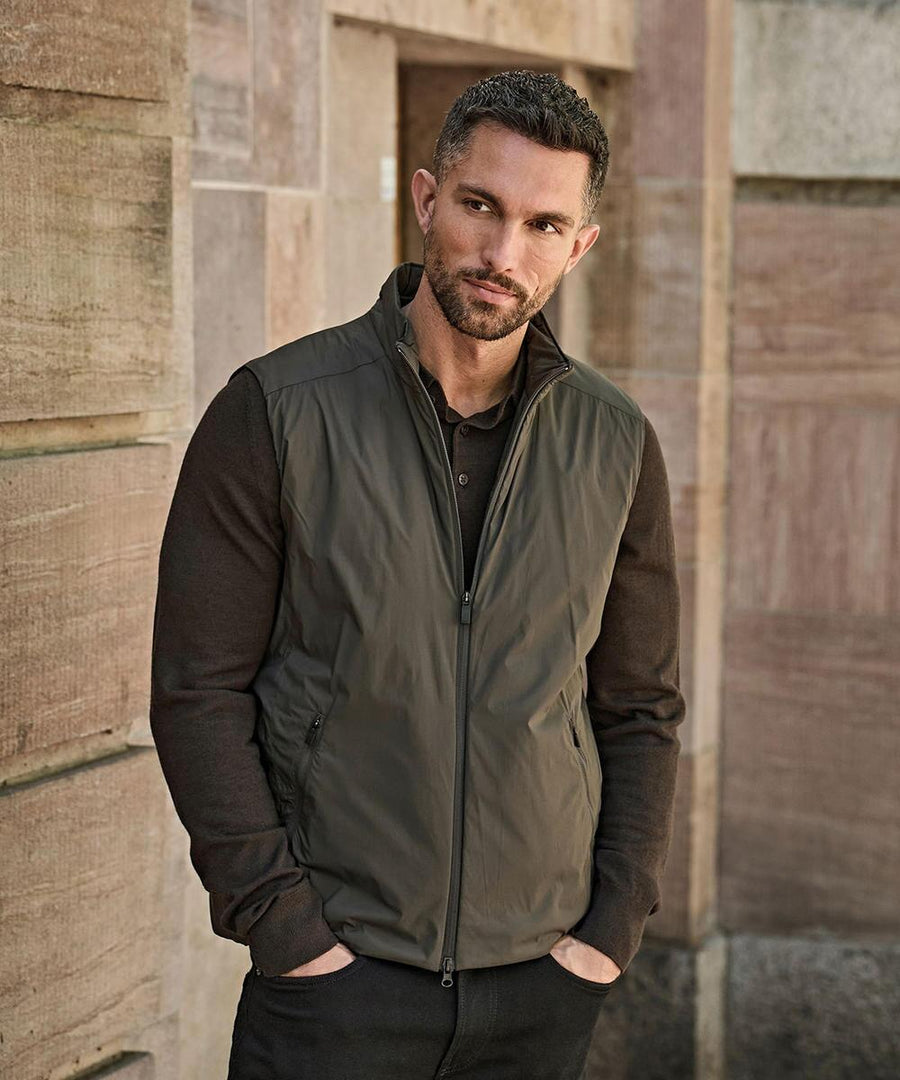 A man with short hair and a trimmed beard stands outside against a stone wall, wearing a Tee Jays PureLite bodywarmer (9692) in olive green over a dark brown long-sleeve shirt, with his hands in his pockets.