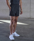 A person wearing a dark shirt, black shorts, and white Tombo TL080 athletic shoes stands on a concrete floor with one hand in their pocket.