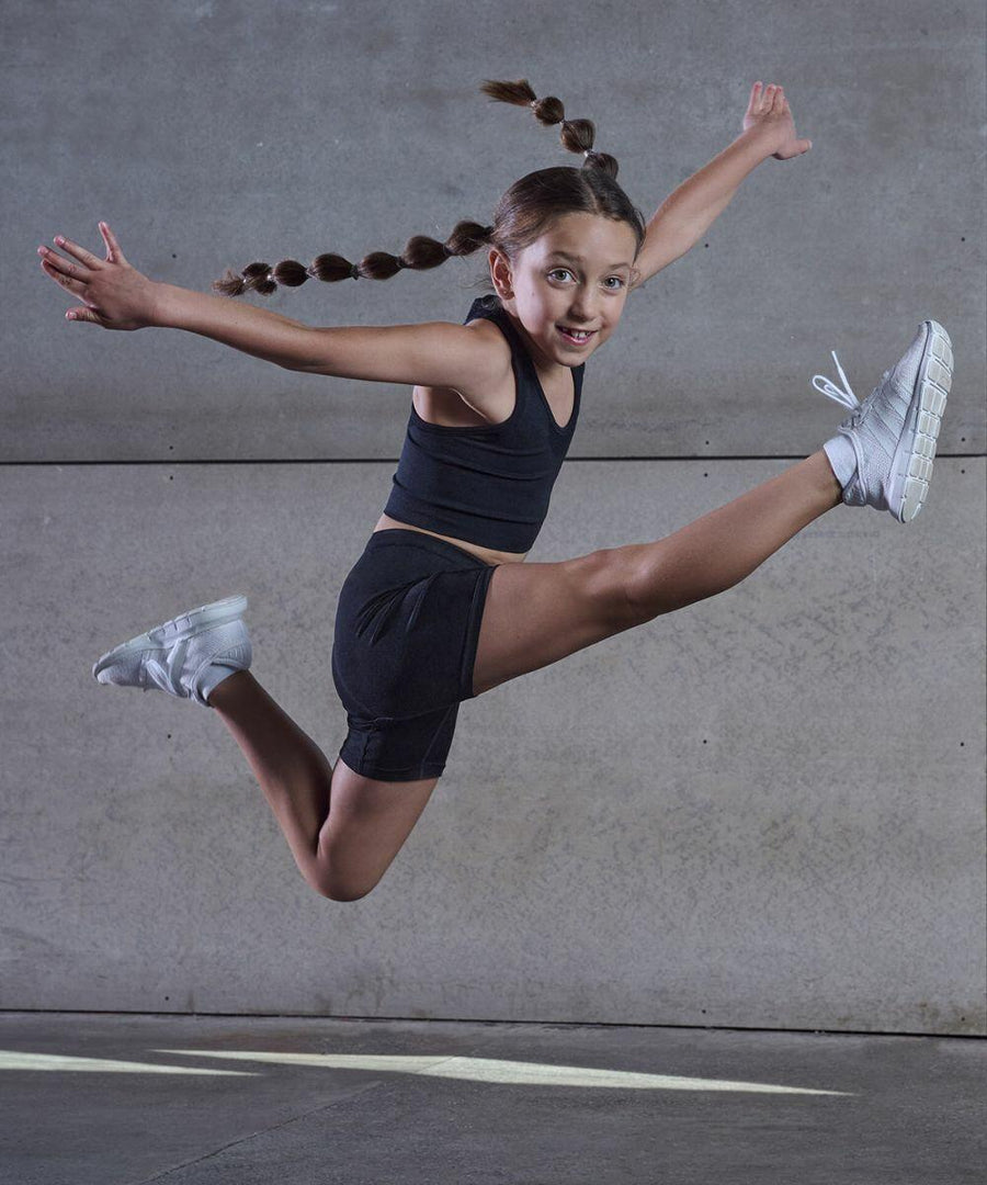 A young girl with braided hair is mid-jump against a concrete wall, wearing a black athletic outfit and white TL309 sneakers by Tombo, her arms and legs extended outward.