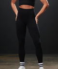 A person stands indoors with hands on hips, modelling Tombo Women's sculpting leggings and a matching black sports bra, paired with white trainers and striped socks. Their head is out of frame.
