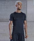 A person with short, bleached hair wearing a black Tombo TL515 athletic shirt and shorts stands in front of a concrete wall, looking to the side.