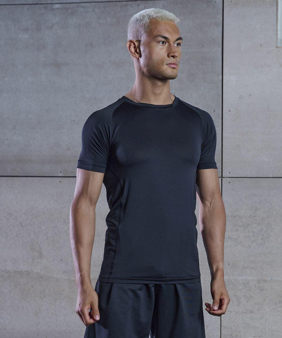 A person with short, bleached hair wearing a black Tombo TL515 athletic shirt and shorts stands in front of a concrete wall, looking to the side.