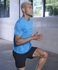 A person with short blonde hair wearing a blue shirt and black shorts is exercising indoors, performing a high-knee march using the TL545 by Tombo.