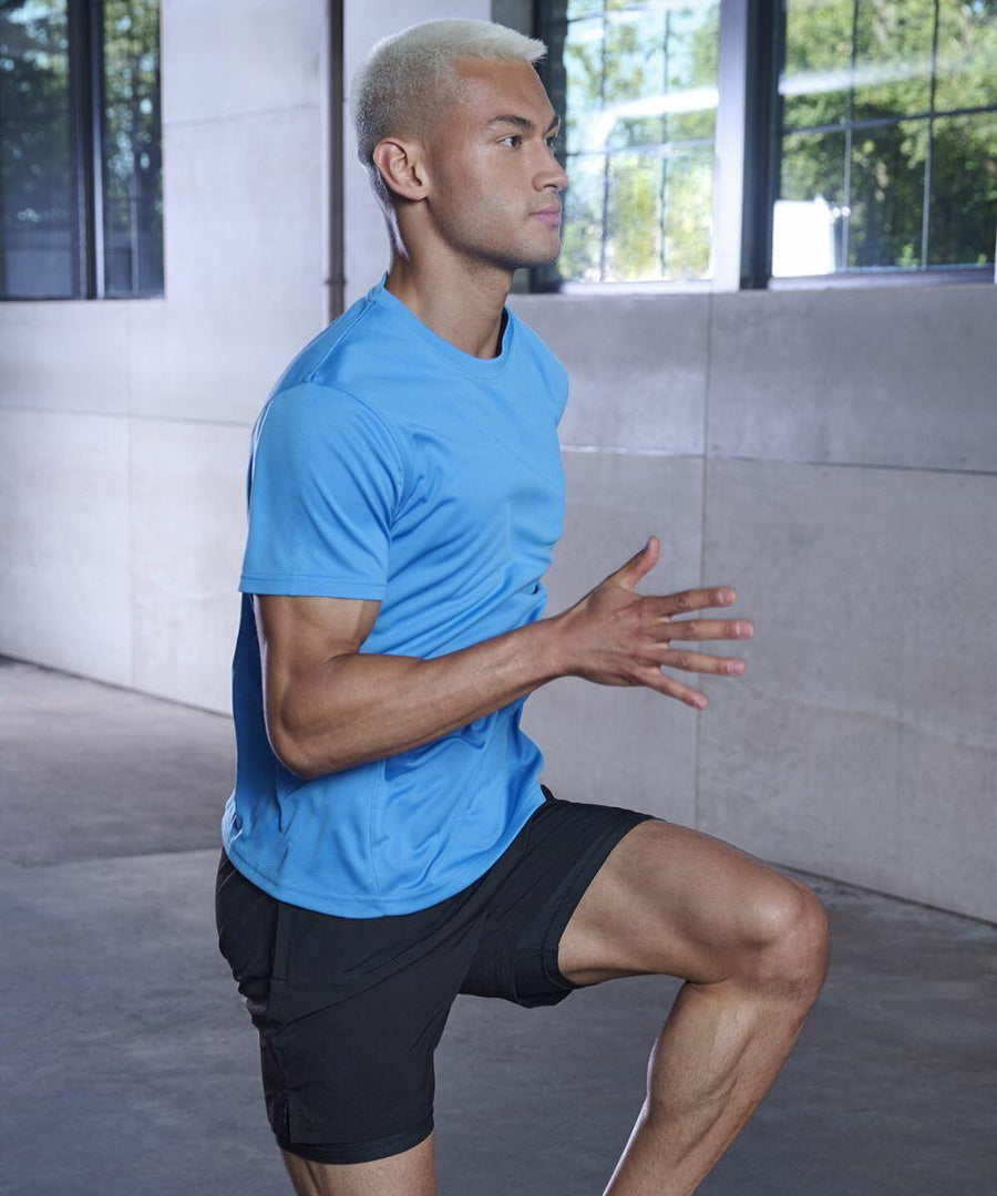 A person with short blonde hair wearing a blue shirt and black shorts is exercising indoors, performing a high-knee march using the TL545 by Tombo.