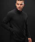 A man with short hair and a trimmed beard wears the Tombo Technical 1/4-zip top in black. He stands against a plain black background, looking left with one hand in his pocket.