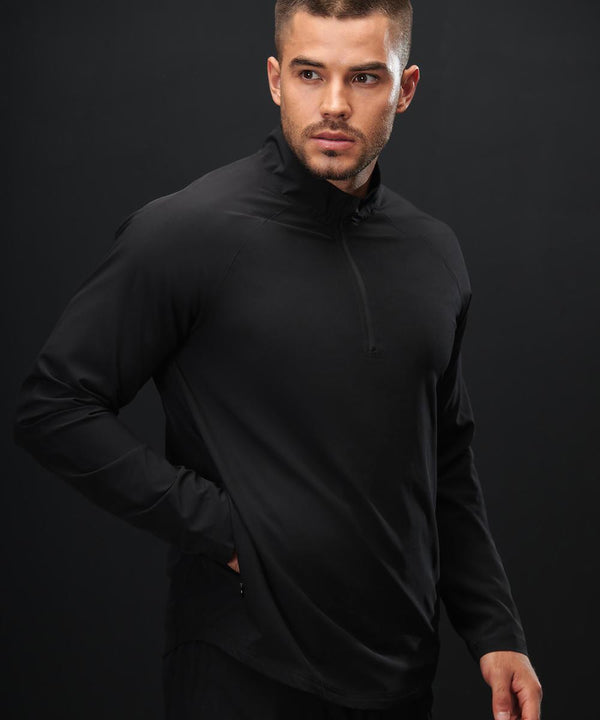 A man with short hair and a trimmed beard wears the Tombo Technical 1/4-zip top in black. He stands against a plain black background, looking left with one hand in his pocket.