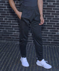 Person standing against a brick wall wearing Tombo TL580 black joggers and white sneakers.
