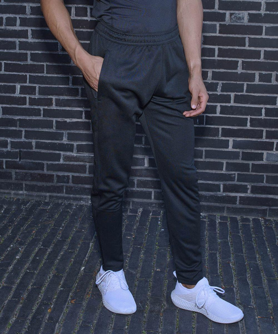Person standing against a brick wall wearing Tombo TL580 black joggers and white sneakers.