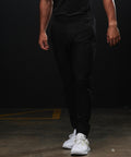 A person stands indoors on a concrete floor, wearing Tombo Technical trousers with a black shirt and white trainers. Their head is out of the frame, and their arms hang relaxed at their sides.