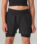 A person wears Tombo Kids double-layer sports shorts in black over black fitted shorts, paired with a black crop top. Their arms are relaxed at their sides against a plain light grey background.