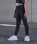 A person wearing a black TL673 sports bra and leggings, and white sneakers stands against a concrete wall. Only the lower half of their body is visible.