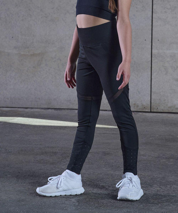 A person wearing a black TL673 sports bra and leggings, and white sneakers stands against a concrete wall. Only the lower half of their body is visible.