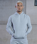Person with short blonde hair stands against a concrete wall wearing a Tombo TL710 light gray hoodie with hands in the front pocket.