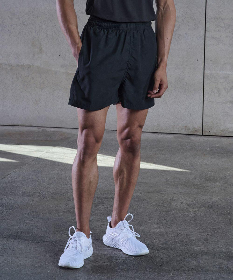 A person wearing black Tombo TL800 athletic shorts, a dark shirt, and white sneakers stands on a concrete floor with a neutral background.