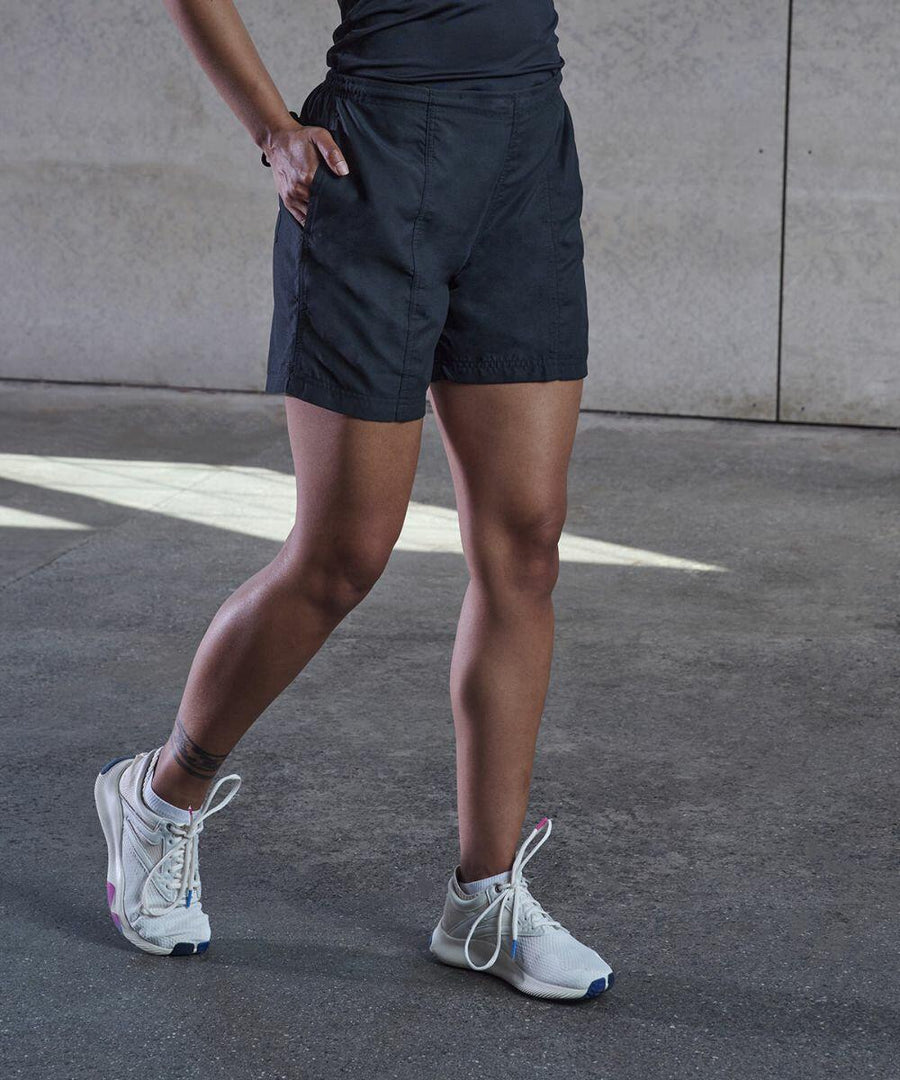 Person wearing black shorts, a black top, and white TL80F sneakers by Tombo, standing on a concrete floor with hands in pockets.