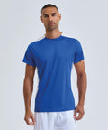 A person stands facing forwards in a TriDri Contrast performance t-shirt by TriDri, featuring blue fabric with white accents on the shoulders and sides, paired with white shorts, set against a plain light background.