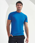 A man in a blue Tri-Dri TR011 short-sleeve shirt and black shorts stands against a light background.