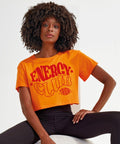 A person with curly hair wearing an orange crop top with "Tri-Dri TR019" written on it, paired with black leggings, poses in front of a white background.