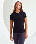 A person with braided hair is wearing a black short-sleeve Tri-Dri TR020 shirt and dark leggings, standing and smiling at the camera against a white background.