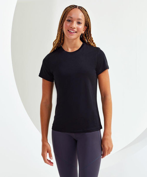 A person with braided hair is wearing a black short-sleeve Tri-Dri TR020 shirt and dark leggings, standing and smiling at the camera against a white background.