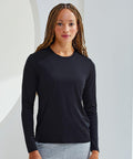Person with long braided hair wearing a black TR060 long-sleeve shirt by Tri-Dri and light gray pants stands against a light-colored background.