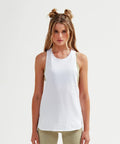 Woman with long hair in a double bun style, wearing a white sleeveless Tri-Dri TR061 top over a beige sports bra and beige leggings, standing against a plain white background, facing forward.