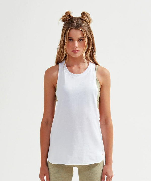 Woman with long hair in a double bun style, wearing a white sleeveless Tri-Dri TR061 top over a beige sports bra and beige leggings, standing against a plain white background, facing forward.
