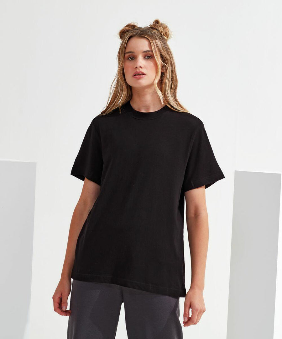 A woman with long hair styled in two buns stands against a white background, wearing a black Tri-Dri TR064 t-shirt and grey pants.
