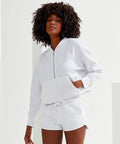 A person with curly hair wears a white Tri-Dri TR077 zip-up hoodie and matching white shorts, posing with hands in the hoodie pocket against a plain light background.