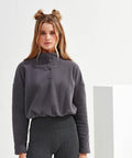 A woman with long hair in two buns is wearing a dark grey Tri-Dri TR087 pullover and dark patterned leggings, standing against a plain white background.