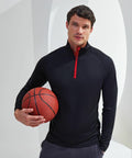 A person in a black long-sleeve Tri-Dri TR110 sports shirt holds a basketball with one hand, standing against a minimalist background.