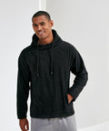 A person wearing a black TR114 hoodie by Tri-Dri and gray pants is smiling and posing with one hand in their pocket against a plain background.