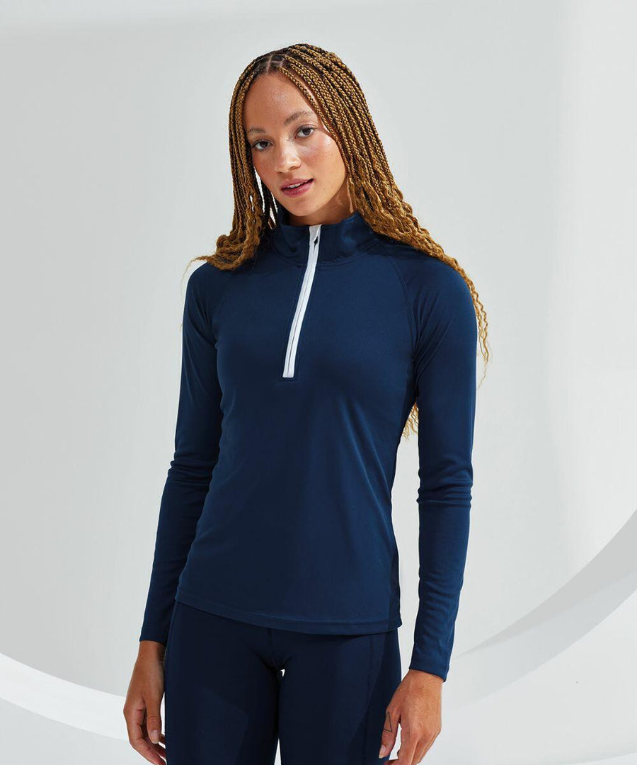 A person with long braided hair is wearing a Tri-Dri TR120 navy blue long-sleeve athletic top with a half-zipper front. They are standing in front of a plain, light-colored background.