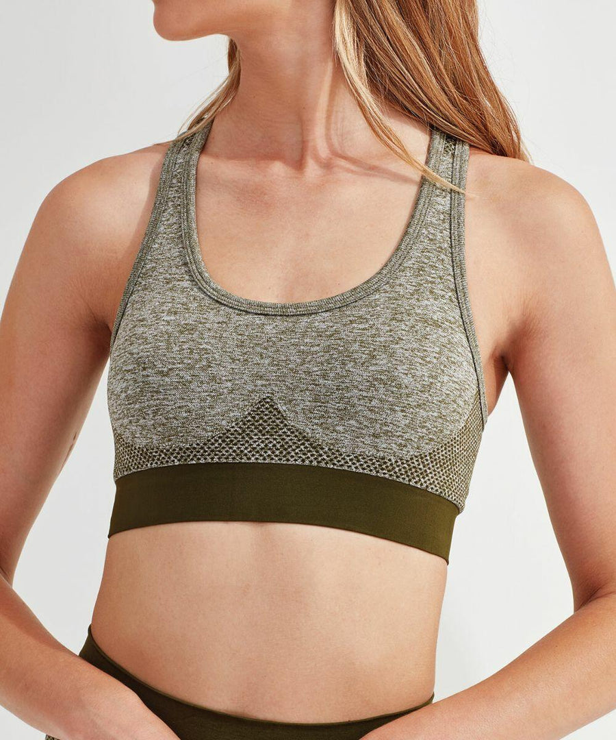 A woman wearing a gray Tri-Dri TR210 sports bra with a green band and green leggings is standing and looking to the side.