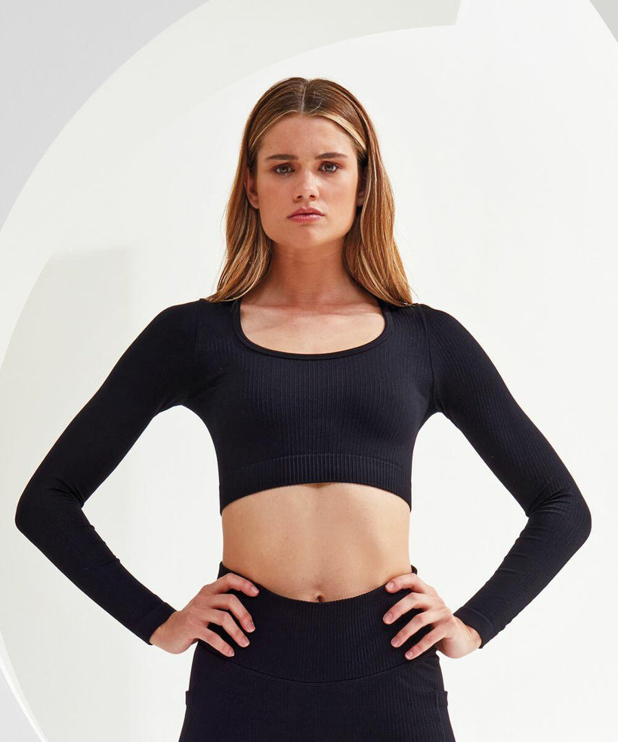 A woman with long hair stands with hands on hips, wearing a black Tri-Dri TR224 long-sleeve crop top and matching bottoms, looking directly at the camera.