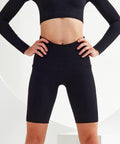Person in a black workout outfit, consisting of a Tri-Dri TR225 long-sleeve crop top and high-waisted shorts, standing with hands on hips.
