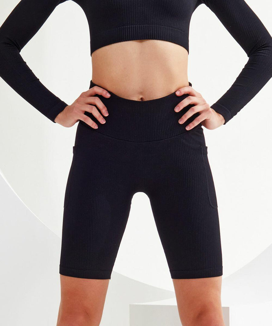 Person in a black workout outfit, consisting of a Tri-Dri TR225 long-sleeve crop top and high-waisted shorts, standing with hands on hips.