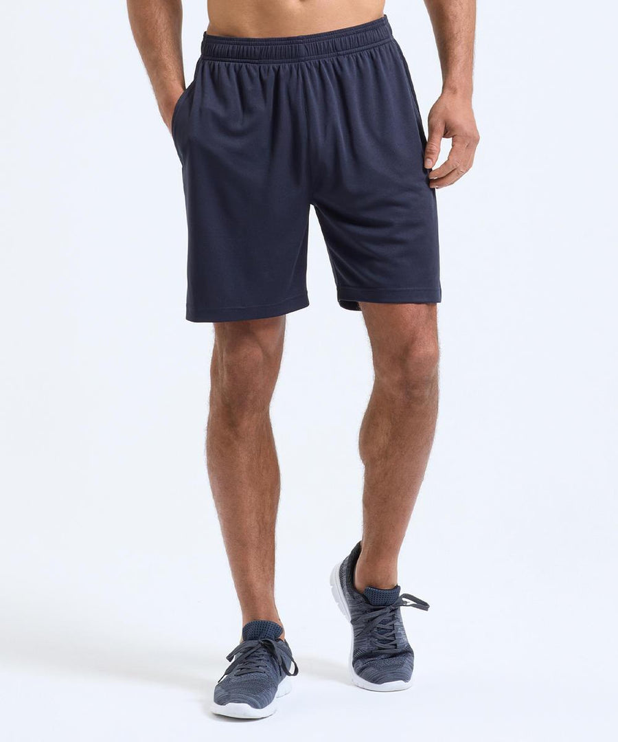 A person wears TriDri Unisex TriDri Ion-Charge performance shorts in navy, with matching navy trainers featuring white soles, standing against a light background. Their hands are in the pockets and their face is not visible.