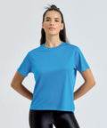 A person stands against a plain light background, wearing the TriDri Women's TriDri Ion-Charge Easy Fit tee in bright blue with black shiny leggings, hands on hips and hair partially tied up.