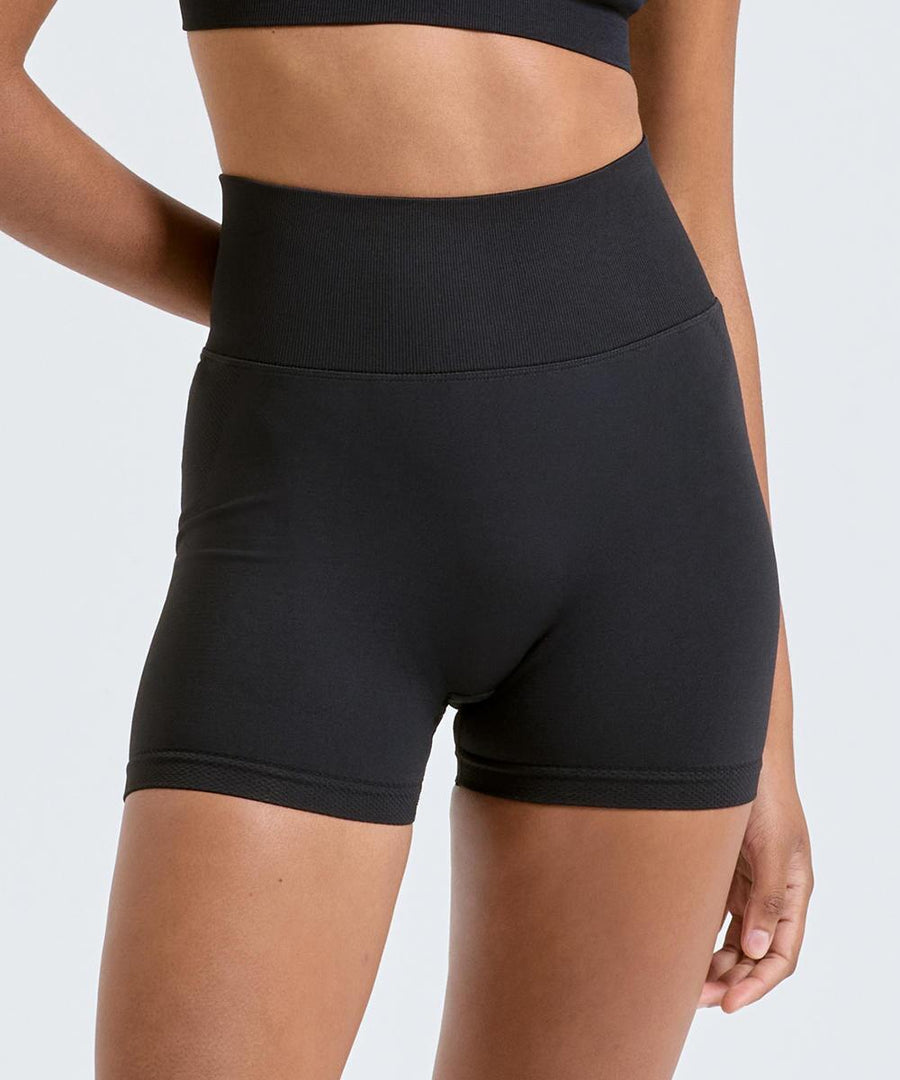 Model wears TriDri's Women's TriDri Scrunch shorts in black with a matching black sports bra, standing against a light grey background. The image is cropped from midsection to upper thigh.