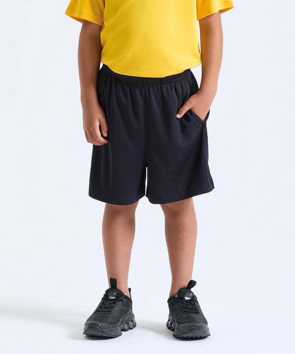 A child models TriDri’s Kids TriDri Ion-Charge performance shorts in black, paired with a yellow shirt and black trainers. The child stands against a plain white background with hands in the pockets; face not visible.