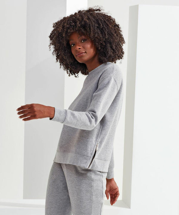 A person with curly hair wearing a grey TriDri TR600 sweatshirt and sweatpants stands indoors against a white backdrop.